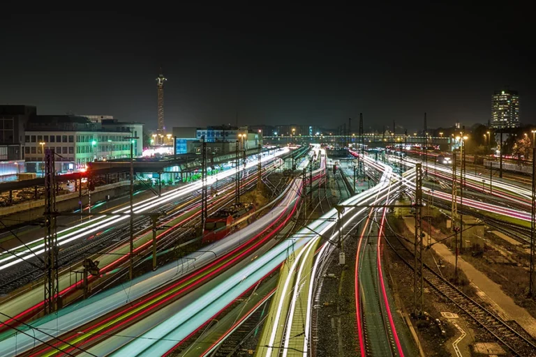 Blick auf Schienenetz und Bahnhof bei Nacht. Durch Langzeitbelichtung ergeben sich bunte Lichtstrahlen der fahrenden Züge